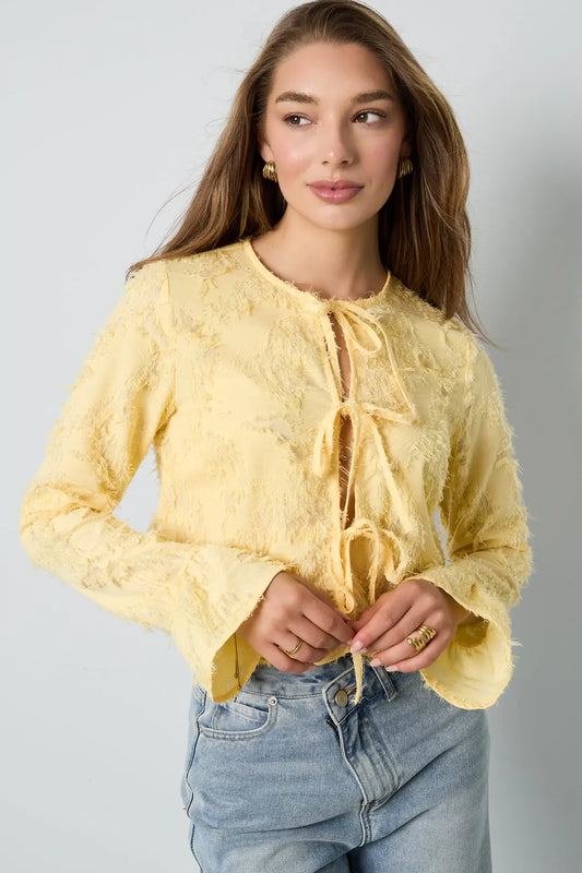 Aanfoto voorkant lichtgele blouse met strikjes en textuur. Lange mouwen met licht uitlopende mouwen.